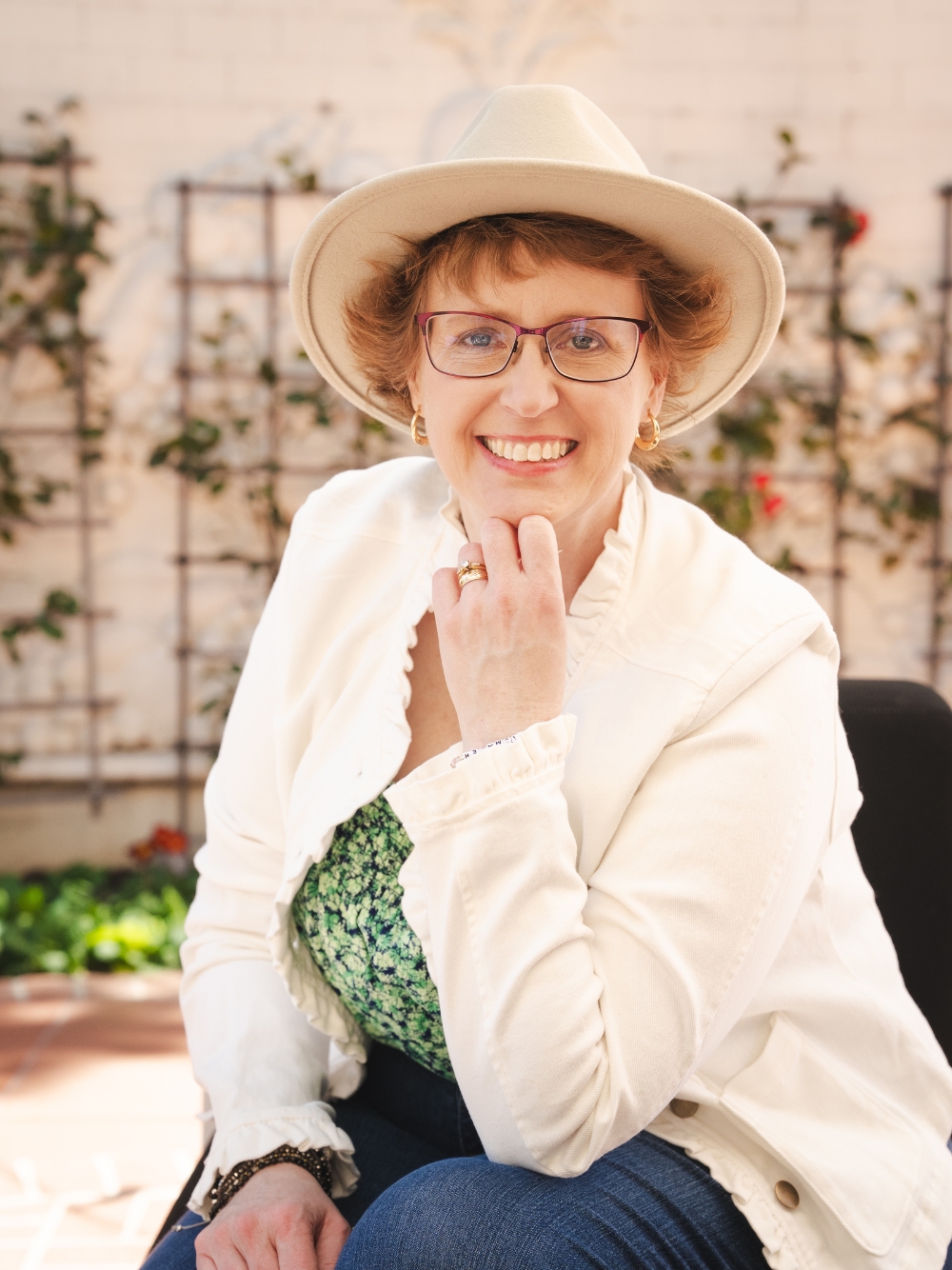 A white woman with short reddish hair smiling at the camera, wearing glasses, a light-colored hat, a cream jacket, and a green floral top, seated outdoors in front of a trellis wall with greenery. Contact Virginia Greer Coaching